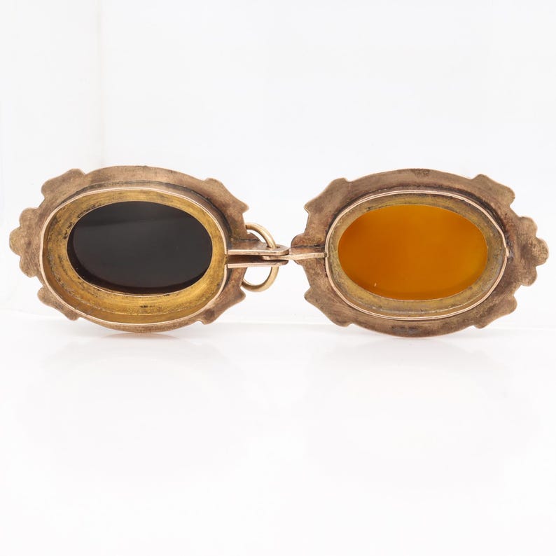 May include: A gold locket pendant with two oval compartments. One compartment has a black background and the other has a yellow background.
