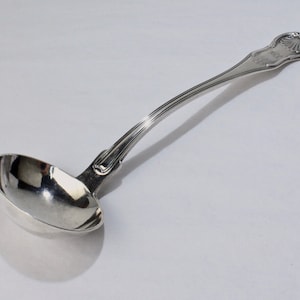 Könnte beinhalten: Eine silberne Schöpfkelle mit einem dekorativen Griff, der ein Muscheldesign aufweist. Die Kelle ist poliert und glänzend.