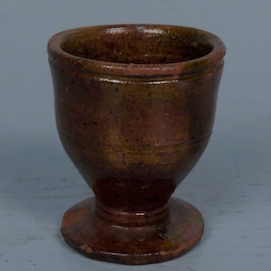 May include: A small, brown ceramic cup with a flared base and a rounded top. The cup has a mottled glaze with shades of brown, green, and orange.