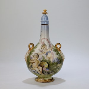 May include: A ceramic bottle with a blue and white lid, two handles, and a painted scene of cherubs in a garden. The bottle is decorated with a light blue, white, and brown glaze.