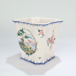 Könnte beinhalten: Ein weißer Keramik-Pflanztopf mit einem blauen und grünen Blumendesign. Der Topf hat eine quadratische Form mit abgerundeten Ecken und einem gewellten Rand.