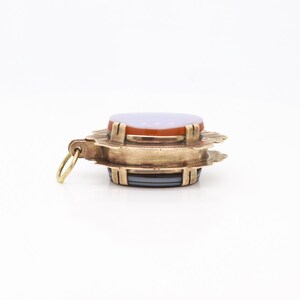 May include: A gold pendant with a layered agate stone. The stone is a mix of purple, orange, and gray. The pendant has a decorative, scalloped edge.