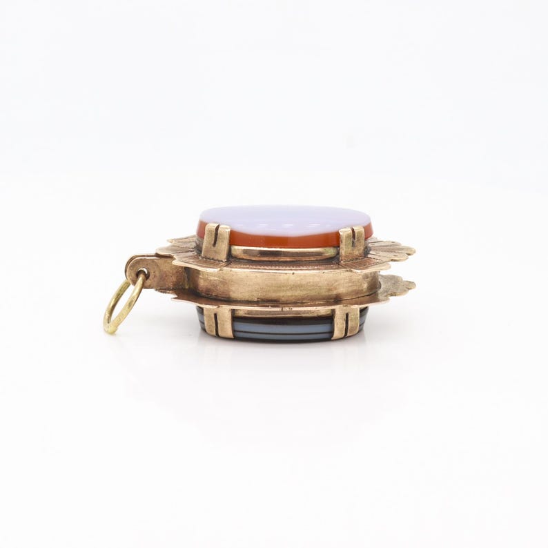 May include: A gold pendant with a layered agate stone. The stone is a mix of purple, orange, and gray. The pendant has a decorative, scalloped edge.