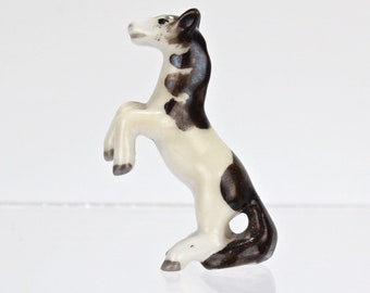 May include: A small ceramic figurine of a black and white horse rearing up on its hind legs. The horse is painted in a realistic style with brown and white markings.