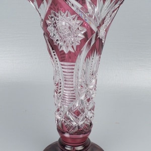 May include: A ruby-colored crystal vase with a flared top and a round base. The vase features intricate cut glass patterns, including starburst and diamond designs. The rim has a scalloped edge, and the vase is approximately 10 inches tall.