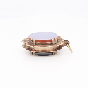 May include: A gold-toned pendant with a layered design featuring a light purple oval stone surrounded by a red oval stone and a decorative gold frame. The pendant has a loop for attaching a chain.