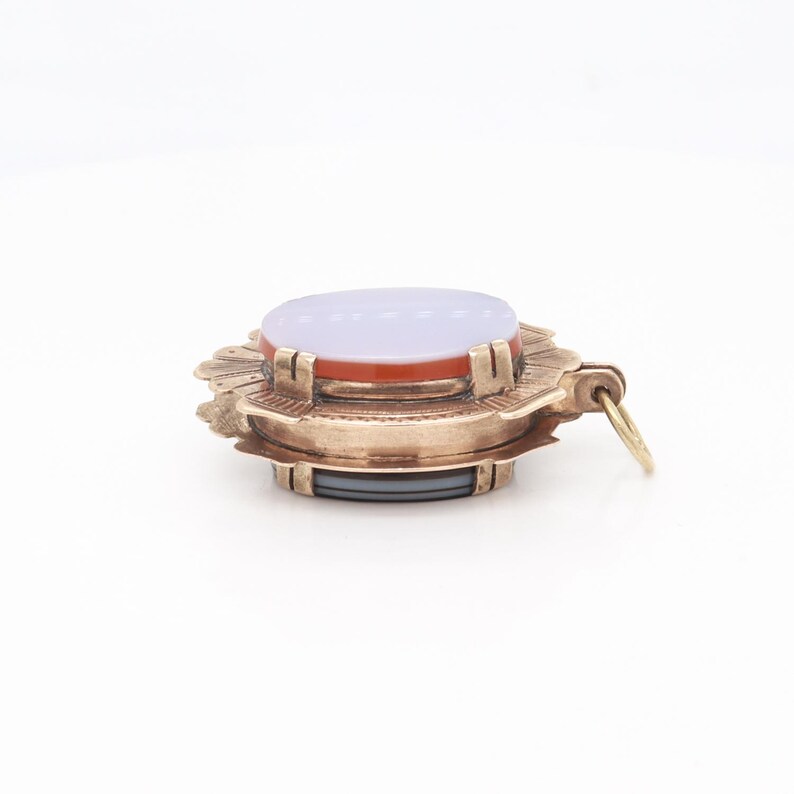 May include: A gold-toned pendant with a layered design featuring a light purple oval stone surrounded by a red oval stone and a decorative gold frame. The pendant has a loop for attaching a chain.