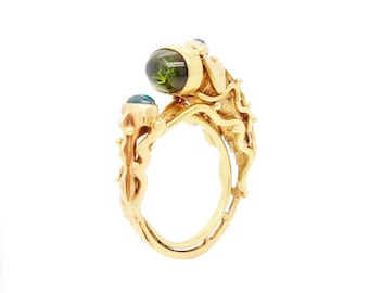 Puede incluir: Un anillo de oro con una piedra preciosa verde y piedras preciosas azules. El anillo está intrincadamente diseñado con un patrón de remolino.