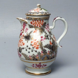 Puede incluir: Una jarra de crema de porcelana blanca con tapa, decorada con un patrón floral en tonos rojos, naranjas y dorados. La jarra tiene un cuerpo redondo, un pico y una asa.