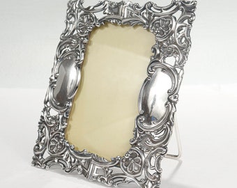 May include: A silver-plated picture frame with an ornate, intricate design. The frame has a stand and is made of metal.