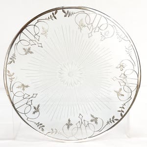 May include: A clear glass plate with a silver rim and decorative etched floral and geometric patterns. The center features a flower design with radiating lines. The plate's edge is adorned with silver leaf and scroll designs.
