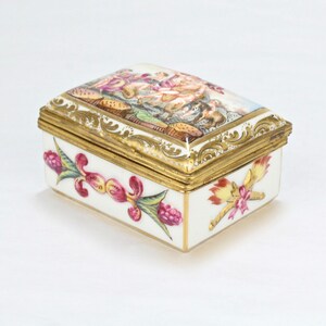 Puede incluir: Una caja de alhajas rectangular de porcelana con una tapa con bisagras. La tapa está decorada con una escena pintada de figuras en un paisaje, enmarcada con detalles dorados. La caja está decorada con motivos florales en rosa, rojo y verde.