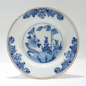 Puede incluir: Un plato de cerámica blanca con un diseño floral y paisajístico azul. El plato presenta una escena central con un árbol y un borde floral. El borde tiene un ribete dorado. Parte del plato está astillado.