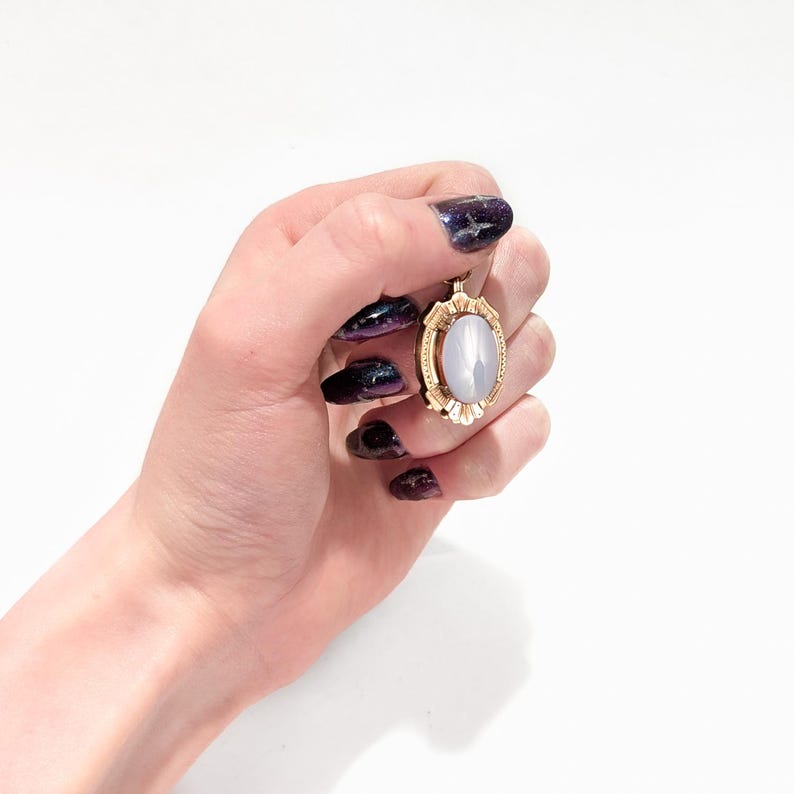 May include: A gold oval pendant with a white stone set in the center. The pendant is surrounded by a decorative gold frame.