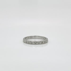 Puede incluir: Un anillo de plata con pequeñas piedras preciosas transparentes engastadas alrededor de la circunferencia. El anillo tiene detalles grabados y marcas en el lateral. El anillo se muestra sobre una superficie blanca.