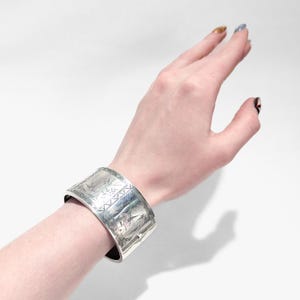 Puede incluir: Un brazalete de plata con un dise&ntilde;o grabado intrincado. El dise&ntilde;o presenta un motivo floral y patrones geom&eacute;tricos.