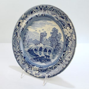 May include: A blue and white ceramic plate with a decorative border and a scene of a bridge and a castle in a landscape.