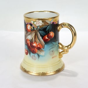 May include: A vintage porcelain mug with a gold rim and handle. The mug features a hand-painted design of cherries on a branch with green leaves. The mug is decorated with gold accents and has the inscription "J.E. Roy" on the bottom.