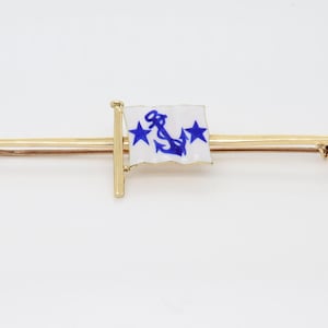 Könnte beinhalten: Goldfarbene Brosche mit weißer Flagge, blauem Anker und Sternen-Design. Die Flagge ist an einer goldenen Stange befestigt, und die Brosche hat eine lange, schlanke Nadel. Das Design ist wahrscheinlich maritim.