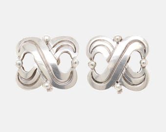 May include: A pair of silver earrings. Each earring has an intertwined, infinity-like design with rounded edges and small, spherical accents. The earrings have a polished finish and a symmetrical shape.