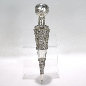 Puede incluir: Un frasco de perfume plateado con un diseño decorativo y ornamentado. El frasco tiene una parte superior bulbosa, una sección media detallada con figuras y una base puntiaguda. La botella de vidrio es visible a través de la ornamentada metalurgia.