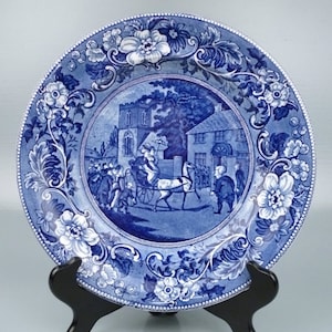 Könnte beinhalten: Blaue und weiße Keramikplatte mit floralem Rand und einem zentralen Bild einer Pferdekutsche vor einer Kirche und Häusern.