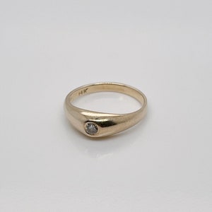 Puede incluir: Un anillo de oro con un solo diamante redondo engastado en el centro. El anillo está marcado con "14K" en el interior.