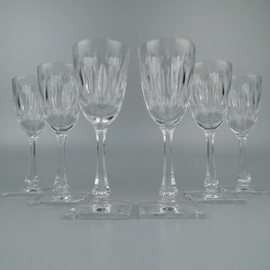 May include: Six clear glass stemmed drinking glasses with a cut glass design. The glasses are arranged in a row on a white surface.