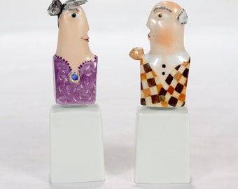 May include: Two ceramic figurines of a man and woman, each standing on a white pedestal. The woman is wearing a purple dress with a blue jewel and has gray hair. The man is wearing a brown and white checkered vest and has gray hair.
