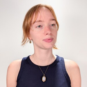 May include: A woman wearing a dark blue tank top with a gold chain necklace and a pendant featuring a white oval stone in a gold frame.