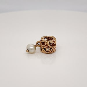 Puede incluir: Un charm de oro con una perla que cuelga. El charm está intrincadamente diseñado con un diamante en el centro.
