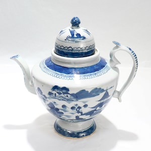 Puede incluir: Una tetera de porcelana blanca y azul con tapa. La tetera presenta un diseño de paisaje con árboles, edificios y barcos. El asa y el pico son curvos. La tapa tiene un pequeño pomo.