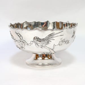 May include: A silver-coloured decorative bowl with a scalloped rim and a pedestal base. The bowl features an embossed design of a bird perched on a flowering branch. The bowl is likely made of metal.