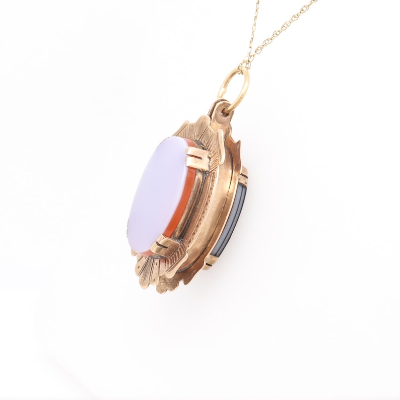 May include: A gold pendant necklace with a cameo featuring a white oval stone surrounded by a red and black stone border. The pendant is attached to a delicate gold chain.