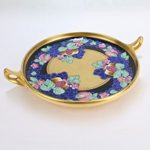 May include: A round serving dish with two handles, featuring a gold rim and a black background with a gold circle in the centre. The circle is surrounded by a blue and green floral design with pink flowers and grapes.