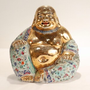 Peut inclure: Statue de Bouddha en céramique dorée, avec un visage souriant et un grand ventre. Le Bouddha est assis, une jambe croisée, vêtu d'une robe à motifs floraux bleus, rouges et verts.