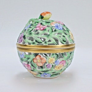 Puede incluir: Una caja de porcelana blanca con un delicado diseño floral. La caja es redonda y tiene una tapa con un pequeño pájaro como adorno. La caja está decorada con hojas verdes y flores de colores.