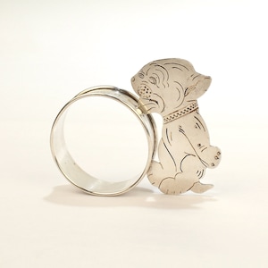 Pode incluir: Anel de guardanapo de prata com um design detalhado de um cão sentado em suas patas traseiras. O anel está aberto e pronto para segurar um guardanapo.