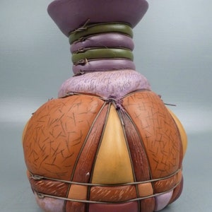 Puede incluir: Un jarrón de cerámica con un cuerpo redondeado y segmentado y una parte superior ancha y plana. El jarrón está decorado con un diseño de patchwork de tonos marrones, naranjas y morados, y está acentuado con finas bandas de alambre marrón.