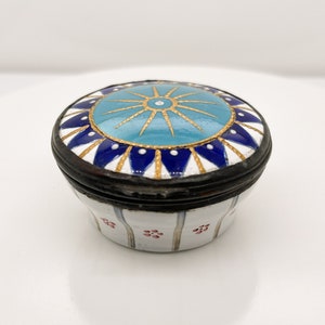 May include: A small, round, white enamel trinket box with a blue and gold lid. The lid features a starburst design with a blue centre and gold rays. The body of the box is decorated with red and white flowers.
