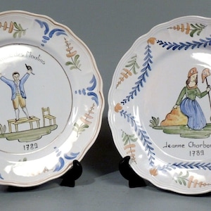 Puede incluir: Dos platos de cerámica blanca con diseños florales azules y verdes. Los platos presentan retratos pintados a mano de personas con los nombres "Charles Noulin 1789" y "Jeanne Charbonnier 1789" escritos debajo.