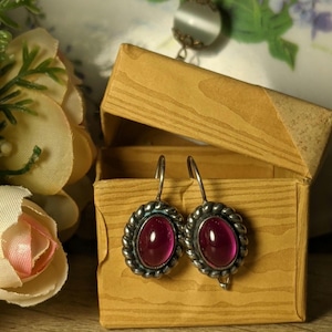 Può includere: Un paio di orecchini d'argento con una pietra ovale rosa scuro incastonata in una montatura d'argento. Gli orecchini sono in una piccola scatola di legno.