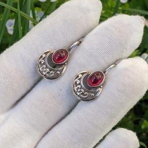 May include: A pair of silver earrings with a crescent moon design. Each earring has a red oval stone set within a floral pattern. The earrings are displayed on a white surface, with green grass and small white flowers in the background.
