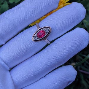 May include: A vintage ring with a red oval gemstone, set in a gold and silver band. The ring is displayed on a white glove, with a yellow flower and green foliage in the background. The ring's design includes two smaller, clear stones on either side of the red gemstone.
