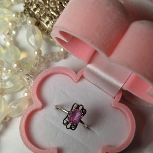 May include: A silver ring with a pink oval gemstone set in a delicate filigree design. The ring is displayed in a pink velvet ring box.