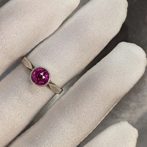 May include: A silver ring with a round, vibrant pink gemstone. The ring is set on a light-colored fabric background. The gemstone is the focal point, showcasing its color and shape. The ring's band is simple and elegant.