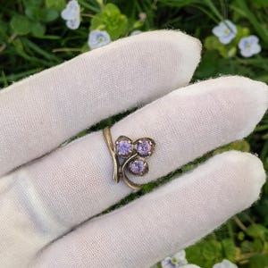 May include: A gold-coloured ring with three light purple gemstones in a cluster design. The ring is set against a white background, with a blurred backdrop of green foliage and small blue flowers.