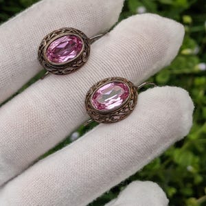 May include: A pair of vintage silver earrings. Each earring features an oval, light pink gemstone set within an ornate, filigree-style frame. The earrings are displayed on a white surface against a blurred green background.