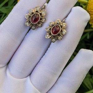 May include: A pair of gold-toned earrings with a flower design. Each earring features a central oval-shaped, deep red stone surrounded by small, clear gemstones. The earrings are set against a white background.