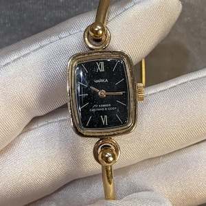 Può includere: Orologio da polso vintage color oro con quadrante nero rettangolare. L'orologio presenta numeri romani per le ore e la scritta "ЧАЙКА" sul quadrante. Il cinturino è in stile bracciale color oro.